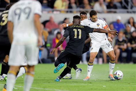 Write informative laconic excerpt under 30 words in mexican spanish for news below. Do not wrap it into quotation marks or html tags. Kei Kamara and Denis Bouanga scored four minutes apart late in the first half to lead LAFC past the LA Galaxy 2-1 on Thursday night in front of 70,076 at the Rose Bowl in a matchup of the top two teams in the Western Conference.It was the second-largest crowd in MLS this season, surpassed only by the 72,610 for Inter Miami&apos;s 3-2 victory over Sporting Kansas City on April 13 at Arrowhead Stadium.Last year&apos;s game drew an MLS-record 82,110 to the Rose Bowl, which was the Galaxy&apos;s home for their first seven seasons (1996-2002).Gabriel Pec scored for the Galaxy, who saw their four-game winning streak snapped. The Galaxy(11-4-7) and Real Salt Lake both have 40 points, but RSL is in second place due to a better goal differential.LAFC has gone unbeaten in its last 10 MLS matches (9-0-1) to move into sole possession of first place in the Western Conference with 43 points (13-4-4).LAFC players celebrate after scoring a goal against the LA Galaxy in MLS.Galaxy goalkeeper John McCarthy made saves on shots by Mateusz Bogusz and Ilie Sánchez in the 39th minute before LAFC converted one minute later on Kamara&apos;s header from a corner kick by Bogusz.It was Kamara&apos;s 147th regular-season goal in MLS, second in league history, while Bogusz has had a goal or assist in 10 straight games.Kamara nearly scored again on a header in the 77th minute, but McCarthy made a diving stop to keep it within one goal.Bouanga scored his 14th goal of the season in the 44th minute when he converted a penalty shot. LAFC was awarded the penalty shot when video review confirmed Galaxy defender Julian Aude tripped Eduard Atuesta just inside the penalty area.LAFC hadn&apos;t lost an MLS contest since May 12, a span of nine matches (8-0-1), coming into the game. In addition to his scoring output, Bouanga leads the club with a career-best nine assists, many of which have set up LAFC&apos;s 2024 breakout star Bogusz, who has contributed to a goal in nine consecutive matches.The Galaxy got on the board in the 56th minute when Pec scored from eight yards out. Miki Yamade was able to get past four LAFC defenders and maintain possession before making the short pass to Pec.After 23 editions of El Tráfico, the series is even at 9-9-5. LAFC has a 49-48 advantage in goals.Information from The Associated Press was used in this report.,Kei Kamara y Denis Bouanga marcaron para LAFC, venciendo al LA Galaxy 2-1 ante 70,076 aficionados en el Rose Bowl. LAFC lidera la Conferencia Oeste.