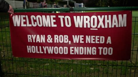 Write informative laconic excerpt under 30 words in mexican spanish for news below. Do not wrap it into quotation marks or html tags. WROXHAM (NOT WREXHAM), England -- Ryan Reynolds and Rob McElhenney stare across the bar in the clubhouse at Wroxham FC. Yet it isn&apos;t the actual Hollywood A-list owners of Wrexham FC, but two life-size cardboard cut-outs of the men who have transformed the Welsh team from non-league no-hopers into one that could reach the Premier League within two years.But this is Wroxham, not Wrexham, and for now at least, the closest that Reynolds and McElhenney will get to this tiny club from an English village on the other side of the U.K. are the cut-outs propped in front of Wroxham&apos;s trophy cabinet. Wrexham or Wroxham? It&apos;s an easy mistake to make, and plenty have done just that."We get calls and things through the [club] website where people have clearly mistaken us for Wrexham," James Blower, Wroxham FC chairman, tells ESPN. "Whether that&apos;s players looking to come and play for us or people wanting the wrong merchandise."We had a commercial partner get very excited when we signed up. I wasn&apos;t initially suspicious because it&apos;s quite usual that you would want to promote a new partnership, but then it became clear when they started talking about doing a viral campaign that they thought they were going to be dealing with Ryan Reynolds. The girls in the marketing team were a little bit disappointed when it was me that they found they were going to be dealing with instead!"Aside from the difference of one vowel -- there are posters in Wroxham about "A team only one vowel away from the big time" -- Wrexham and Wroxham could not be further apart, beyond the 250 miles that separate the Welsh city and the East Anglian village with a recorded population of just 1,502 in the 2021 UK Census.Wrexham are flying high in EFL League One, having secured back-to-back promotions since Reynolds and McElhenney bought the club in February 2021. They are now two promotions away from the Premier League and their story, documented in the hit show "Welcome to Wrexham," has caught the attention of sports fans across the globe.Wroxham are second-bottom of the Isthmian League North, five divisions below Wrexham, and play against teams like Concord Rangers, Brightlingsea Regent and Sporting Bengal United. Tuesday&apos;s home game against Felixstowe & Walton United at the Ginger Pickle Arena, a 2-2 draw, drew an attendance of 221. "We were pleased with that, given the weather," Blower said, noting temperatures had dropped to -10 Celsius [14 Farenheit] with the windchill factor.Wrexham play in red, Wroxham play in blue-and-white stripes and there really is a world of difference between the two teams, but the phone calls from confused football fans continue and bemused visitors still turn up at Wroxham&apos;s ground wondering where on earth they have ended up."It&apos;s happened to me a couple of times," Barry Letten, Wroxham&apos;s 78-year-old groundsman told ESPN. "On one occasion, a person drove here in his car and said, "Is this Wrexham?" "I said, "no, this is Wroxham." And he said, &apos;Oh blimey, I&apos;ve come to the wrong ground!&apos;"So what happened? He stayed, but on another occasion, we had a gentleman that came down and he decided to drive back, I assumed, to Wroxham -- sorry Wrexham! Now I&apos;m getting mixed up!"Wroxham know full well that their story is just as romantic as that of their near-namesakes, but they&apos;re patiently waiting for a shot at the big time. Courtesy of ESPNIt&apos;s not always a mistake when people turn up at Wroxham with Wrexham on their mind. "I&apos;m here on business, so I thought might as well spend a tenner here towards Wroxham&apos;s pockets rather than sitting in the pub drinking," Jim Gregson, a Wrexham fan in his Wrexham shirt, told ESPN during Tuesday&apos;s game."We [Wrexham] have been in the same position as Wroxham. We were on our a-- and got lucky, so if Wroxham want to jump on the back of what we have done, good luck to them."In an attempt to cash-in on their unusual connection with Wrexham -- six letters, basically -- Wroxham have produced their own mini-documentary called "Welcome to Wroxham" and the ambition is to catch the attention of Reynolds and McElhenney, with the ultimate dream of hosting Wrexham in a preseason friendly."That&apos;s been something actually that the Wrexham fans have come and said," Blower said. "There&apos;s been loads saying &apos;We hope every year we draw you in the FA Cup and we want a preseason friendly.&apos; That would be great. But if we&apos;re honest, that wasn&apos;t something we thought of as maybe an outcome from this. I think our purpose is we wanted to showcase what our amazing volunteers do, how great they are for us as a club and highlight the differences between the few levels and how hard it is."Wroxham&apos;s story is one that typifies football at grassroots in the UK. There are so many towns and villages that seem too small to sustain a football club, but ardent supporters, benevolent local businesspeople and selfless volunteers enable clubs to survive even the toughest financially difficulties.Terry Baxter, the club chaplain, once performed the same role for inmates at Norwich Prison. Now in his 80s, he prepares the boardroom on matchdays, laying out sandwiches and arranging blue-and-white teacups in orderly fashion on a freshly ironed tablecloth."I&apos;ve been coming here for 45 years," he said. "I get to plenty of away games too."Groundsman Barry admits he can&apos;t stay away, whether there is a game to be watched or not. "I come here most days," he said. "Today I&apos;ve been doing the car parking, but if there&apos;s a need for some painting and that type of thing or unblocking drains etc, I help out. I&apos;m a general-purpose labourer!"Cardboard cut-outs of Wrexham&apos;s celebrity owners, Ryan Reynolds and Rob McElhenney, have prominent placement in the club&apos;s bar should anyone turn up in vain. Courtesy of Mark Ogden / ESPNIn the bar, Rachel takes care of the catering -- burgers, chips and cheesy fries -- with her teenage children. Before the two sets of players leave after the game, she serves baked potato with beans and cheese for them all in the clubroom."Years and years and years I&apos;ve been here," Rachel said. "It feels like it anyway! It&apos;s my social life actually. I don&apos;t have one outside of here. I&apos;ve always helped with local football."Yet Rachel is less enamoured by the prospect of the club basking in the glow of Wrexham&apos;s celebrity. "I don&apos;t do Hollywood glamour at all," she said. "Definitely not."None of the volunteers are paid for the hours they commit to Wroxham. Only the players and manager earn money from the club, but they all have day jobs for their main source of income. Robbie Linford is a scaffolder, Rio Abodunde works in his family&apos;s restaurant, Harry Barker is a teacher, while goalkeeper Ollie Sutton, the son of former Blackburn, Chelsea and Celtic forward Chris, works in property development."Our players train twice a week and play once or twice a week depending on the fixtures," Blower said. "It&apos;s fairly normal at this level. We&apos;ll play a Tuesday night and they all go and do their day jobs."They have a pretty tough time playing football. We&apos;re the most northerly team in this division, so they have to do a lot of travelling. Sixteen away games in our league are around a hundred miles&apos; away from us, so if this was a Tuesday night away game, the players would be getting back at 1:30 or 2:00 in the morning. So it&apos;s a fair commitment from them."The community spirit of volunteers and part-time players ultimately binds clubs like Wroxham together. Club sponsor Riot Labs even backed an initiative to help Wroxham fans quit smoking, teaming up with Blower to make the Kenny Cooke Stand -- capacity: 50 people -- a smoke-free zone. The hope is that any raised profile from their mini-doc will help make the club ground a more viable community venue."If we could get Wrexham here, we&apos;d fill the ground," Blower said. "We can get 4,000 in here, and that would generate enough for us to clear the debt and buy some furniture for the clubhouse. "That would enable us to make it available beyond matchdays: that kind of thing makes all the difference at this level."The Wroxham fanbase is passionate despite their non-league status. Everyone at the club hopes for a friendly against the Welsh side that could help boost their long-term finances. Courtesy of Mark Ogden / ESPNThe Wrexham dream remains just that, though. Unless Reynolds and McElhenney are drawn in by the romance and quirkiness of a Wroxham vs. Wrexham clash, Wroxham will continue to take the phone calls and redirect any lost visitors.There is one faint glimmer of hope of attracting some of their own Hollywood glamour."So [Hugh Jackman&apos;s] mum is from Norwich [eight miles from Wroxham]," Blower said. The Hollywood A-Lister, who starred alongside Reynolds in "Deadpool & Wolverine", "had the chance to buy into Norwich in 2016. They got promoted straight after that, and he missed out. It&apos;s definitely an opportunity here. If he wants to come and buy in here, I&apos;m happy to take that phone call and we&apos;ll have a conversation."His mum&apos;s still local. We go to the same cinema -- not together -- but yeah, we go to the local cinema. He&apos;s often spotted around here when he is back seeing her and he&apos;s got that friendship with Ryan. So we could definitely have the Vowel Derby with Hugh on one side and Ryan on the other, or Wolverine vs. Deadpool, if you want to call it that."Over to you, Hugh. ,Wroxham, no Wrexham: confusión por clubes de fútbol en Inglaterra. Reynolds y McElhenney propietarios famosos, Wroxham sueña con Premier League. Diferencias entre ambos equipos.